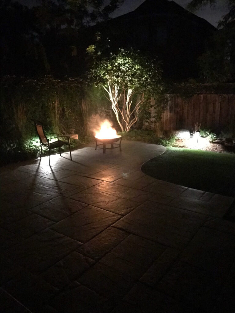 A patio at night with a fire pit and an uplit tree